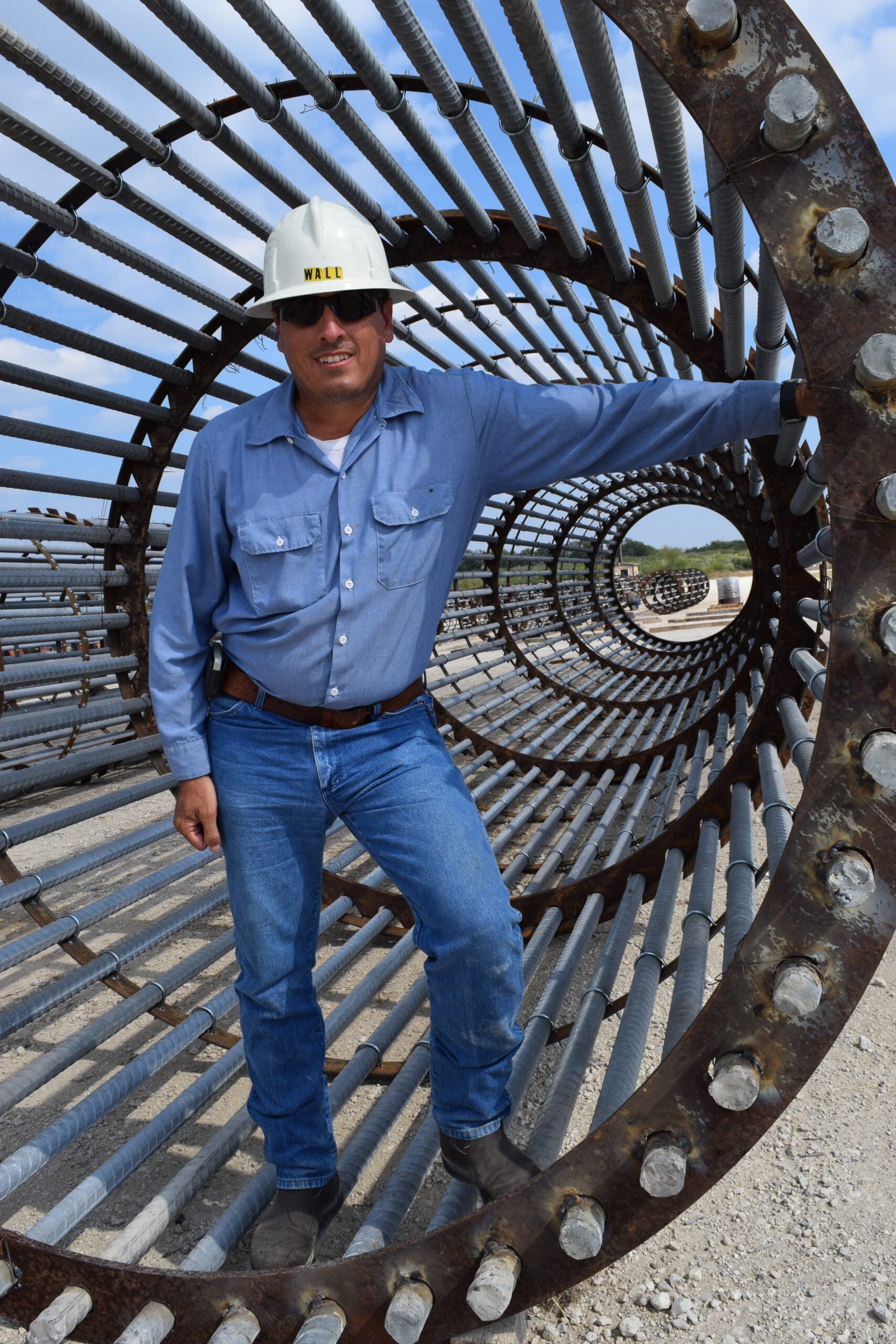 Construction worker poses inside a large metal frame, showcasing industrial materials against a clear sky, emphasizing safety and engineering.