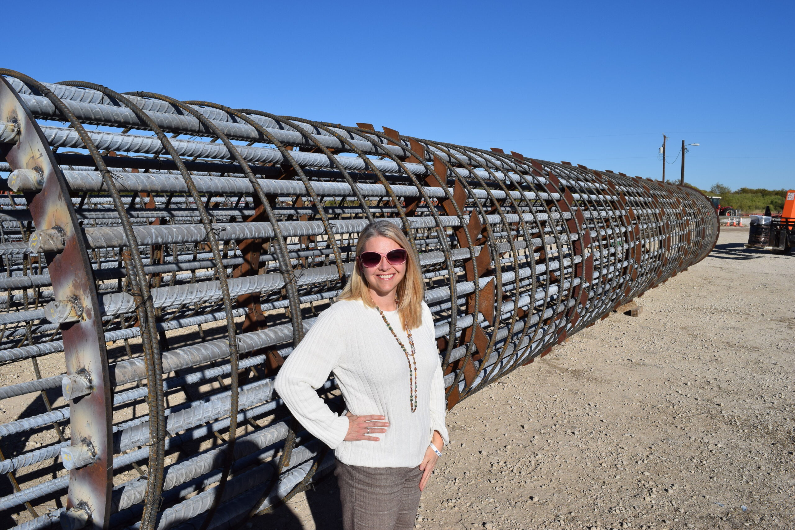 A woman smiles beside a large, cylindrical structure made of metal rods on a construction site, showcasing industrial equipment.