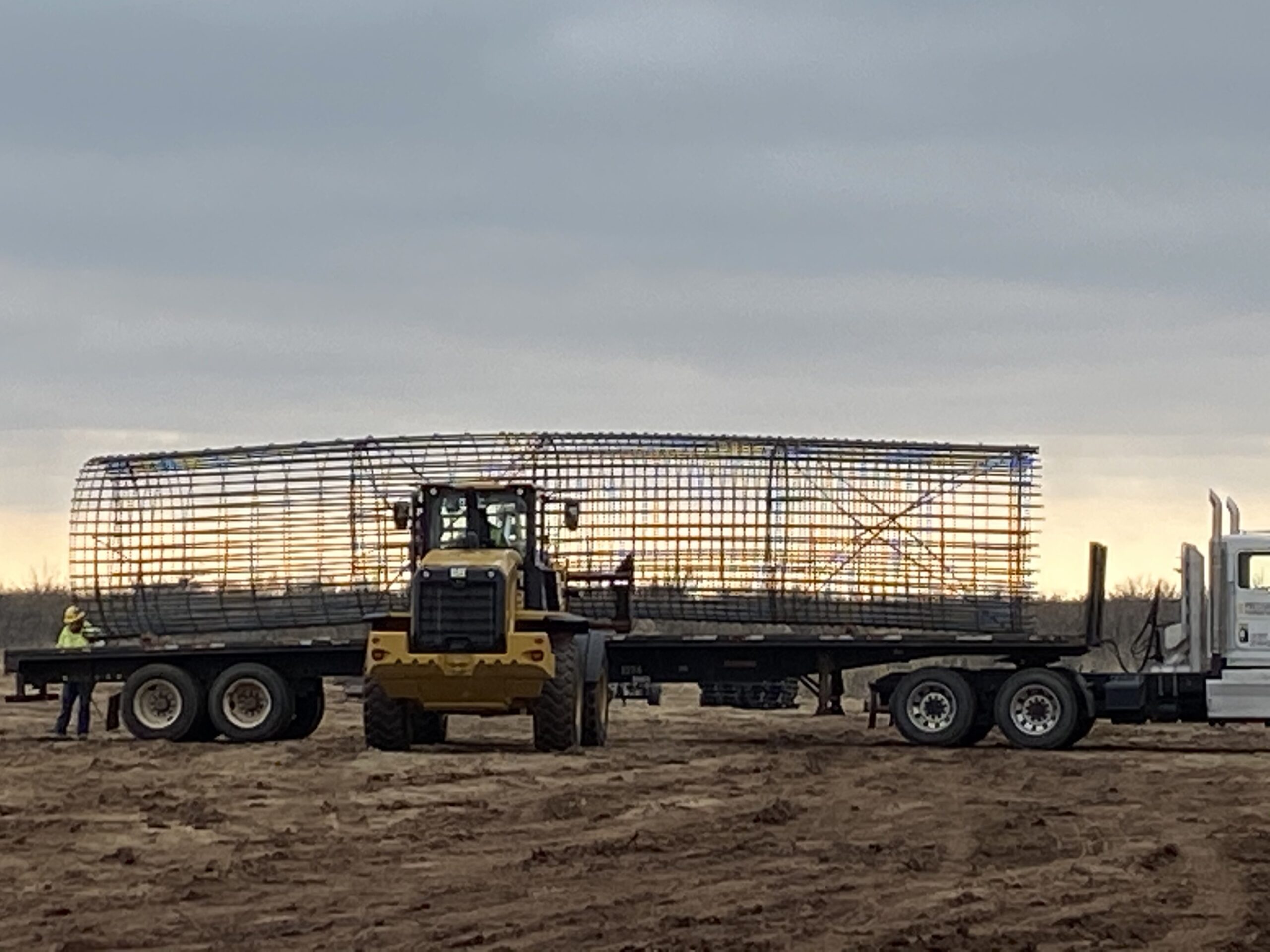 Heavy machinery transports a large metal framework on a flatbed truck across a sandy landscape, marking progress on a construction site.
