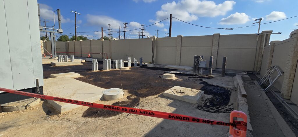 Construction site featuring exposed utility connections, marked by caution tape. Equipment and pipe stubs are arranged on asphalt, indicating ongoing work.