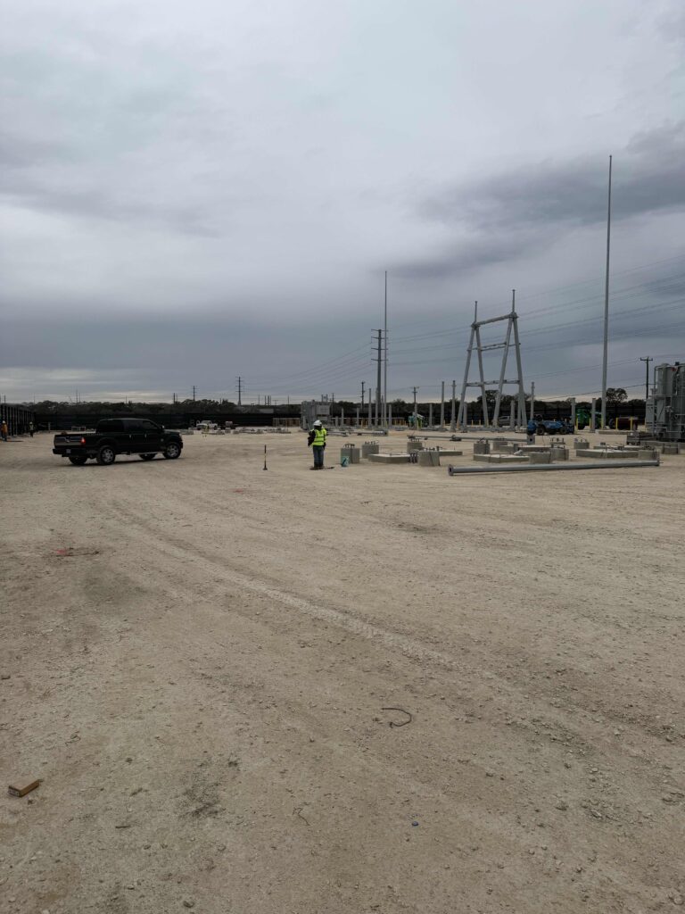 Construction site featuring a worker in a green safety vest, surveying equipment, and concrete structures under gray skies.