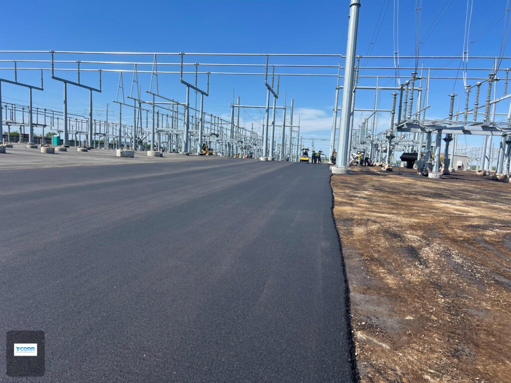 Newly paved asphalt surface at a utility construction site, with electrical infrastructure in the background and workers present, indicating ongoing development.