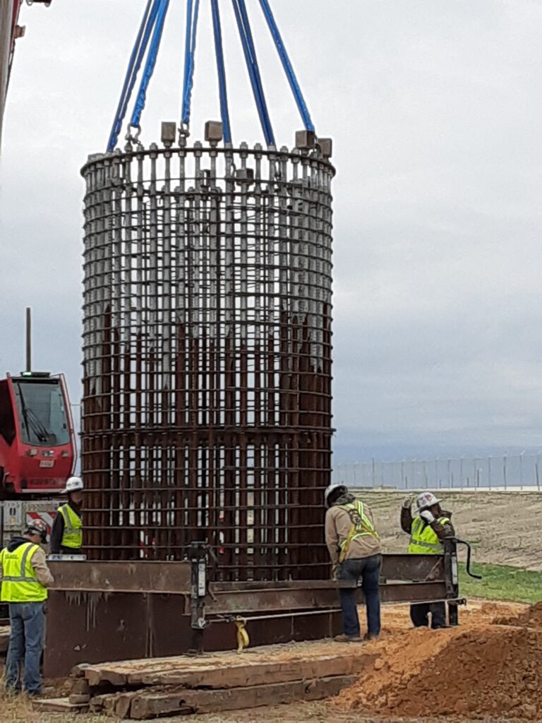 Workers oversee the lifting of a large cylindrical rebar cage at a construction site, preparing for concrete pouring.