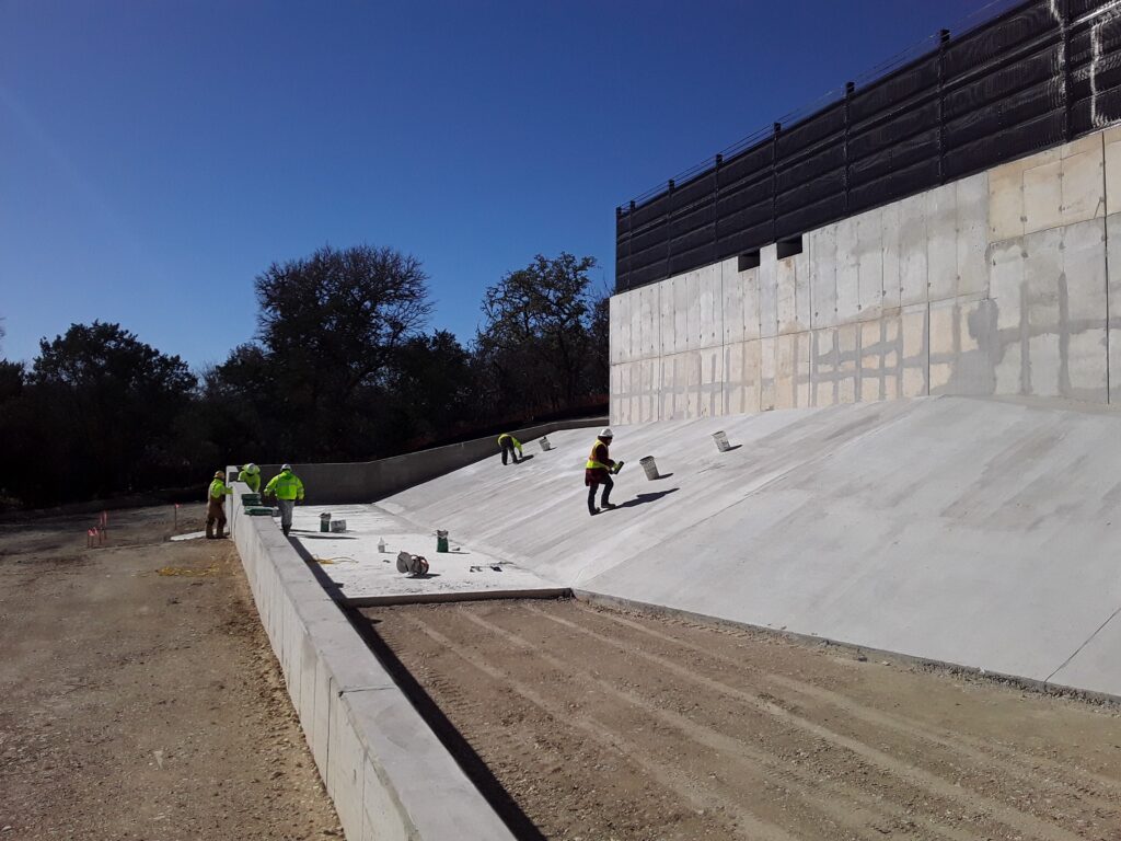 Workers in bright safety gear are constructing a concrete slope, applying finishing touches to a retaining wall in a sunny outdoor setting.